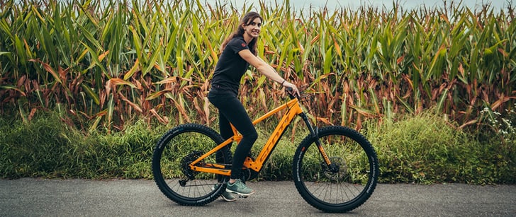 Alexandra, collaboratrice de Bike World, en selle sur son VTT électrique orange flash, devant un champ de maïs.