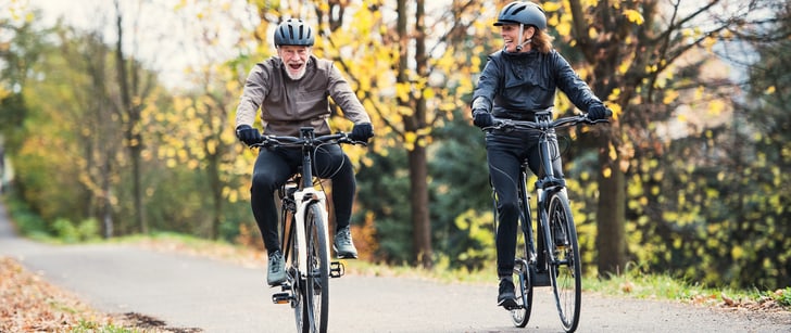 Una coppia di anziani pedala sorridente nel bosco in sella a biciclette elettriche.