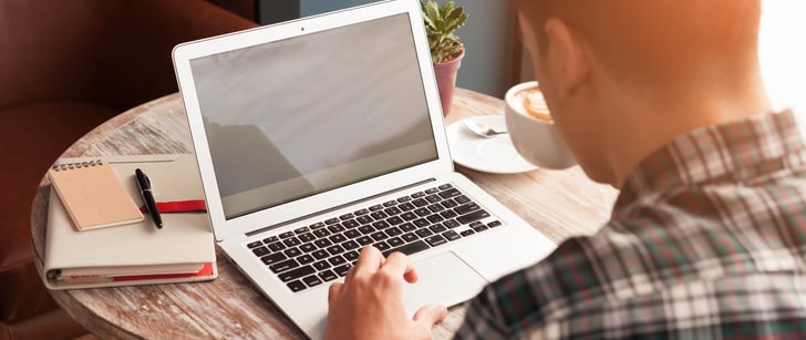 Un uomo seduto a un tavolo rotondo lavora al suo laptop e beve un cappuccino.