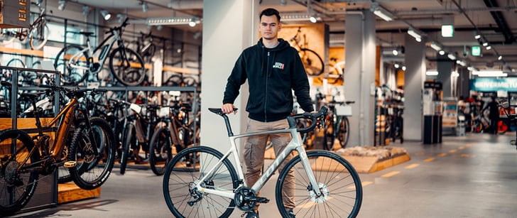 Valmir, collaboratore di Bike World, in posa nel negozio di biciclette dietro alla sua bicicletta da corsa elettrica grigia.