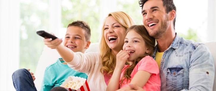 Una famiglia di quattro persone siede sul divano sorridente e guarda insieme un film mangiando popcorn.