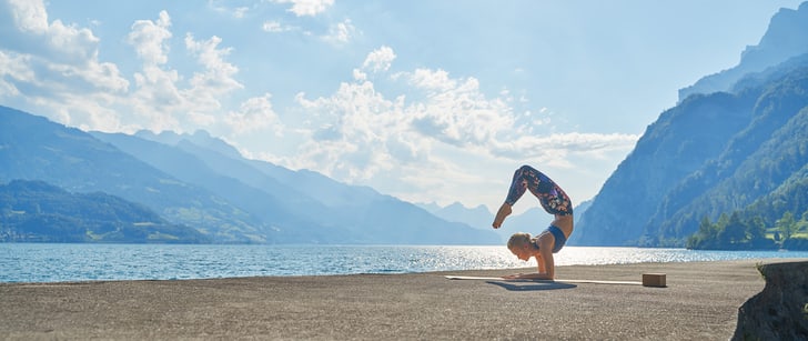 Eine Frau macht eine Yoga-Figur auf ihren Ellenbogen am Seeufer mit einer Berglandschaft im Hintergrund.