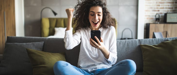 Une femme assise en tailleur sur un canapé regarde l’écran d’un smartphone et lève le poing pour exprimer sa joie.