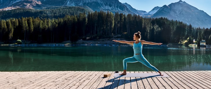 Eine Frau praktiziert Yoga auf einem Holzsteg am See mit Wald und Schneebergen im Hintergrund.