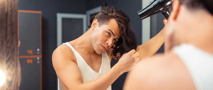 Ein Mann mit Sidecut betrachtet sich im Spiegel während er seine langen Haare föhnt und stylt.