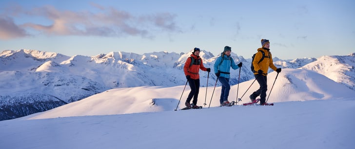 Zwei Frauen und ein Mann in Wintersportausrüstung laufen mit Schneeschuhen durch eine verschneite Berglandschaft.