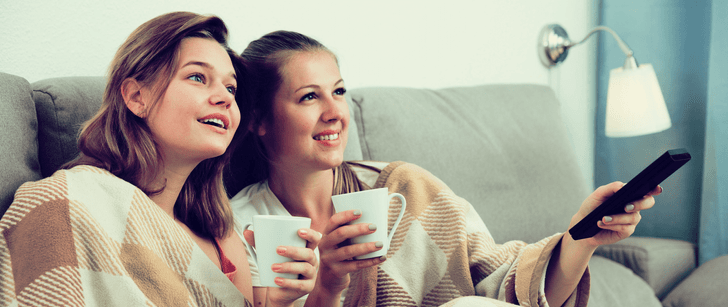 Zwei Freudinnen sitzen auf einem Sofa unter einer Decke und trinken etwas. Dabei schauen sie zusammen einen Film an.