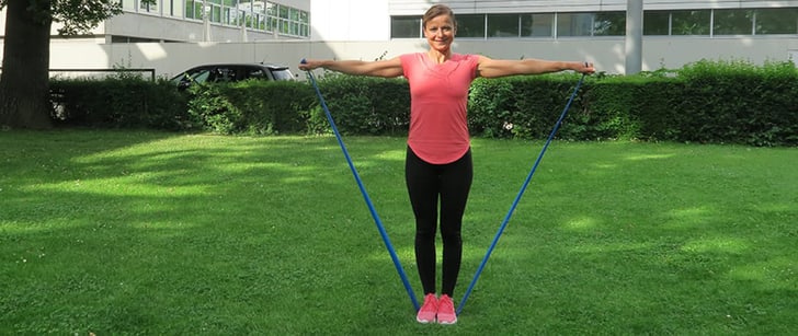 Fabiana montre un exercice de fitness: elle a les deux pieds posés sur une bande élastique et tend les bras.