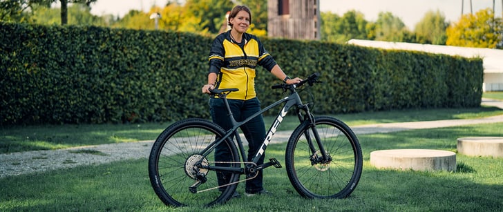 Die Bike World Mitarbeiterin Claudia stützt sich in einem grünen Garten auf ihr schwarzes Cross-Country Bike.