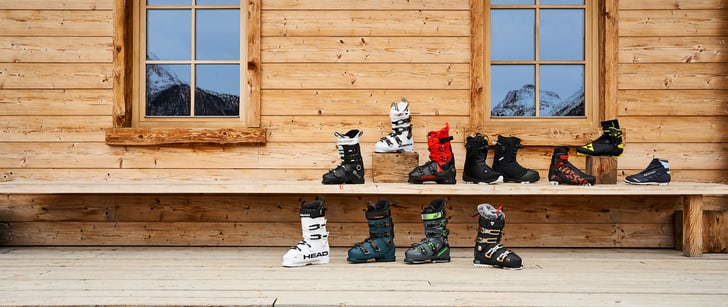 Vari scarponi da sci, scarpe da sci di fondo e scarponi da snowboard appoggiati a una parete di legno chiaro di una capanna.