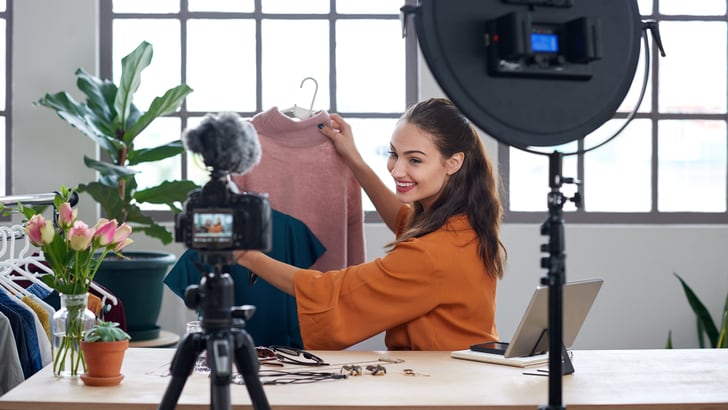 Femme assise présentant deux vêtements à la caméra qu’elle a installée sur une table.