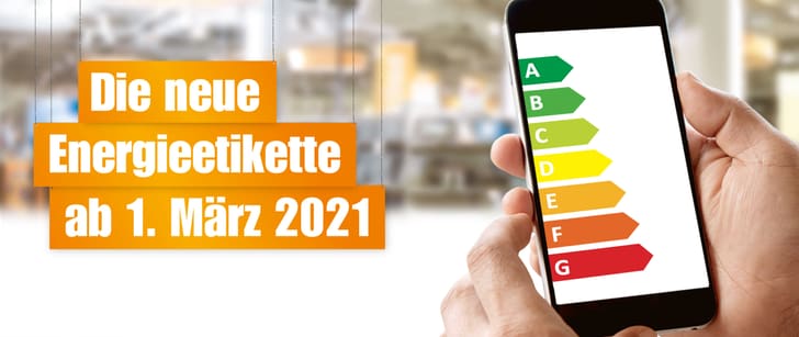 Orange Schilder mit der Aufschrift: “Die neue Energieetikette ab 1. März 2022” hängen von Schnüren. In der rechten Ecke hält eine Hand ein Smartphone auf dessen Bildschirm die neuen Etiketten zu sehen sind.