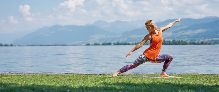 Eine Frau praktiziert Yoga auf einer grünen Wiese am See mit einer Berglandschaft im Hintergrund.