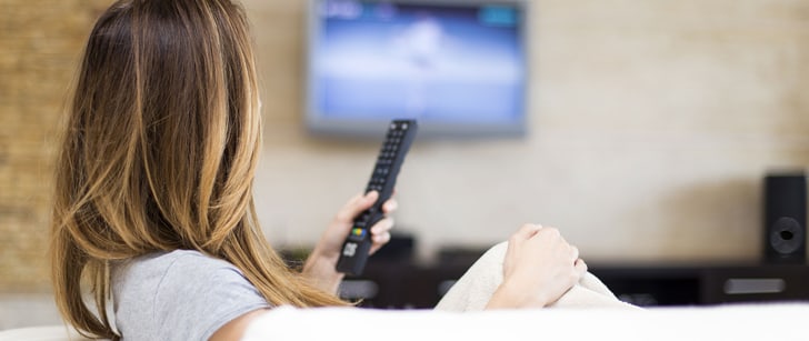Une femme assise dans un canapé tient une télécommande et regarde un écran au mur.