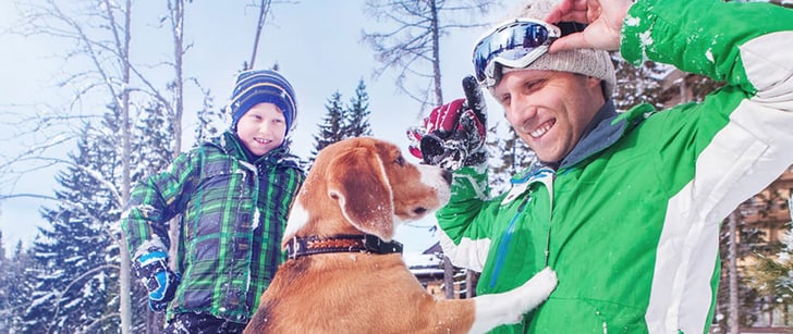 In un paesaggio innevato, un cane mette le zampe anteriori sul petto di un uomo in abiti invernali mentre un ragazzo li osserva.