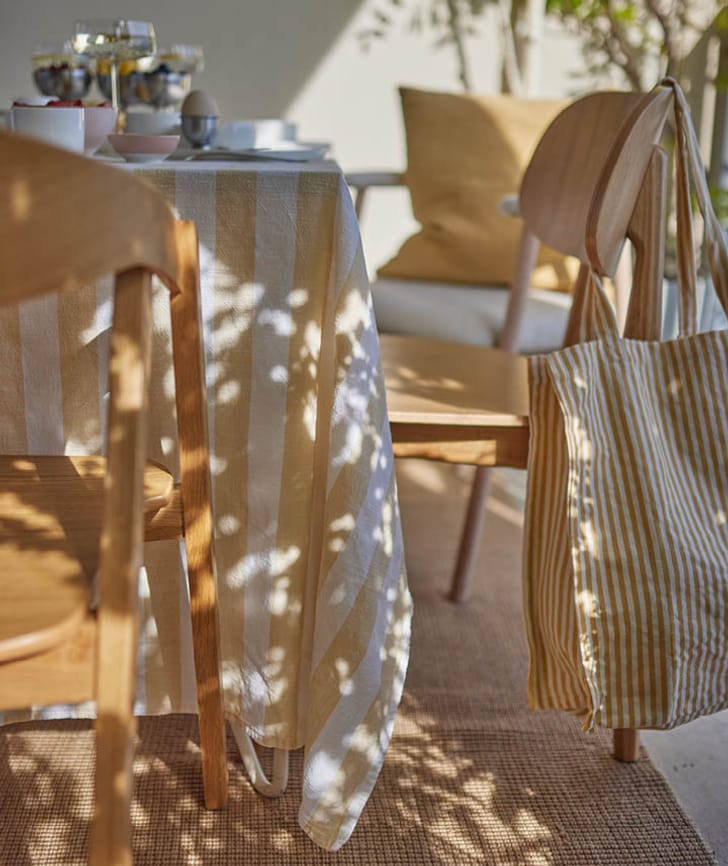 Ein gedeckter Tisch mit gestreifter Tischdecke, Holzstuhl und gestreifter Tasche im Sonnenlicht.