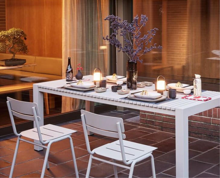 Ein gedeckter Gartentisch mit Geschirr, Gläsern, zwei Laternen und einer Vase mit lila Zweigen, im Hintergrund eine Sitzbank vor einer Fensterfront.