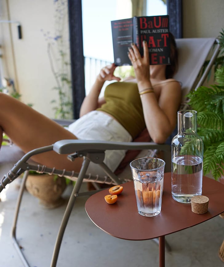 Entspannte Szene auf einem Balkon: Ein Glas Wasser mit Aprikosen, eine Karaffe und eine Person beim Lesen.