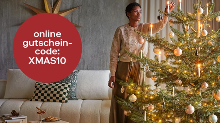 Eine Person dekoriert einen Weihnachtsbaum in einem gemütlichen Wohnzimmer, neben einem roten Kreis mit dem Text &quot;Online Gutschein-Code: XMAS10&quot;