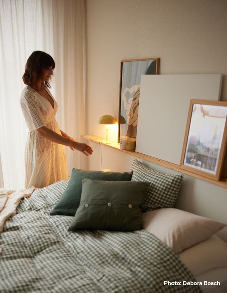 Une personne en robe blanche se tient à côté d&apos;un lit avec une literie à carreaux verts et des coussins verts. Une lampe jaune est allumée.