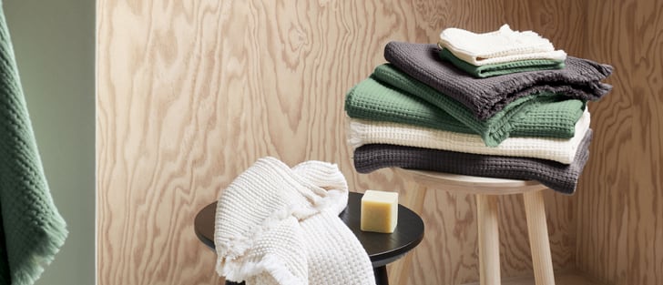 Ein gestapeltes Musselin-Handtuch-Set in Grün, Creme und Grau auf einem Holzhocker. Ein weiteres cremefarbenes Handtuch liegt auf einem schwarzen Hocker.
