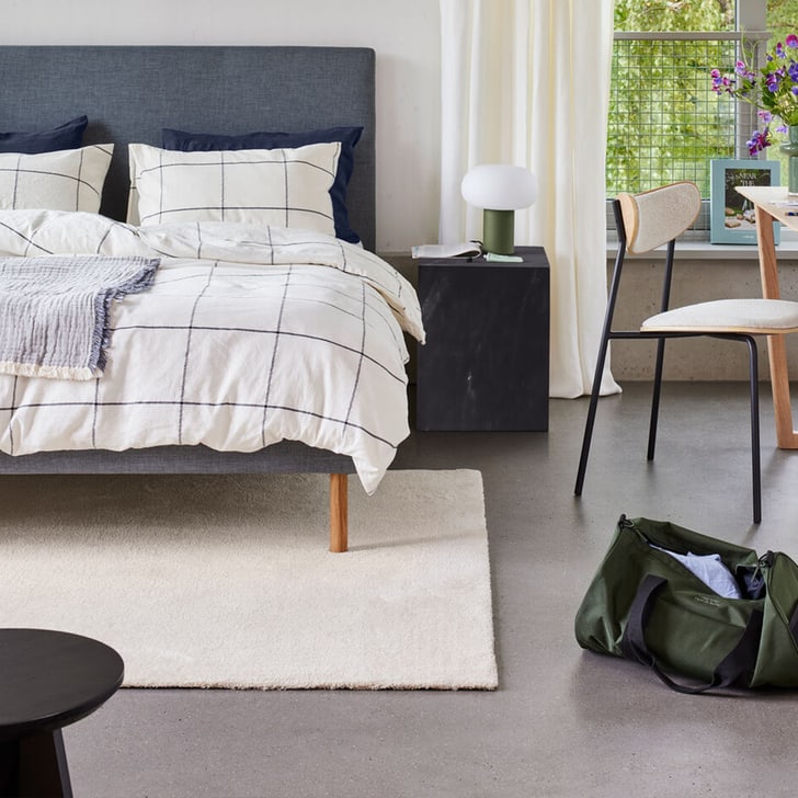 Chambre avec lit gris, linge de lit a carreaux, lampe de chevet et sac a dos vert sur le sol.