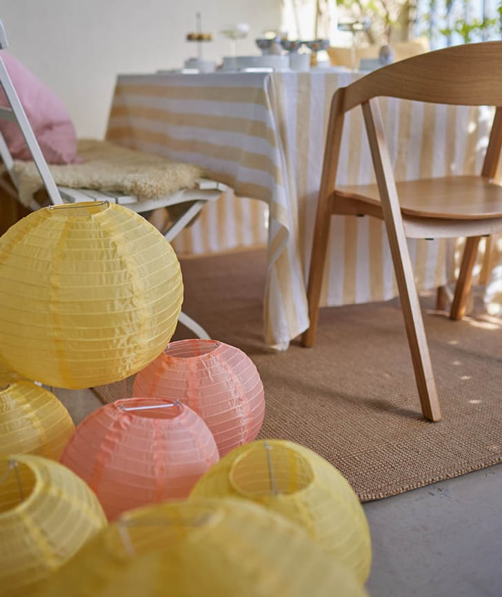 Gelbe und orangefarbene Laternen neben einem Tisch mit gestreifter Tischdecke und Holzstühlen, im Hintergrund Narzissen.