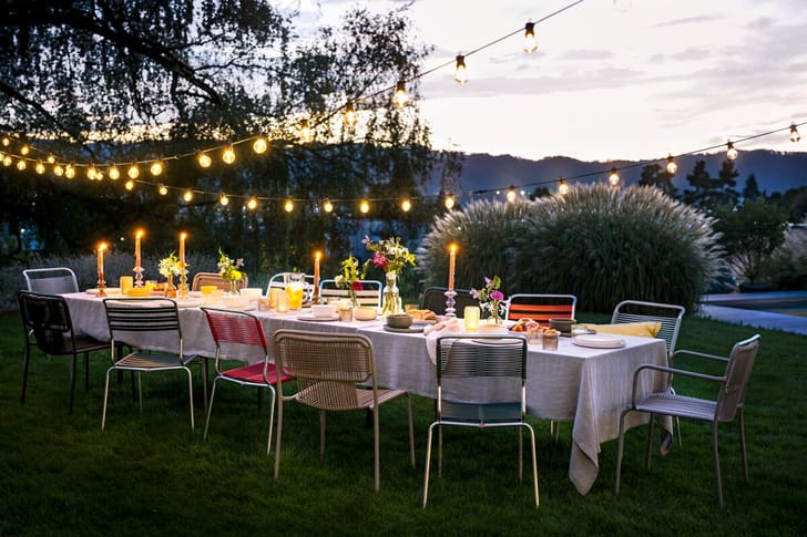 Table de jardin décorée pour une fête, au crépuscule, avec guirlandes lumineuses et bougies.