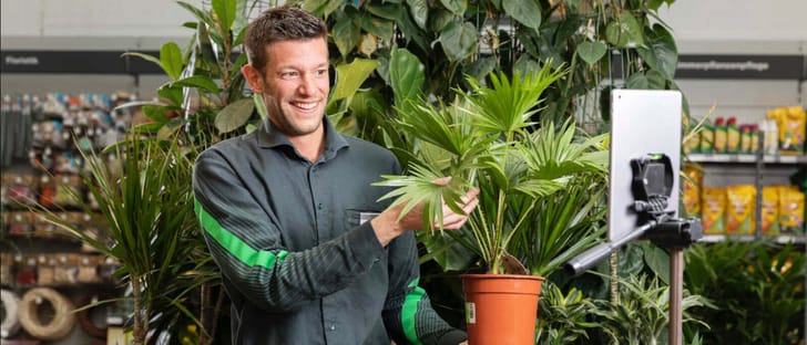 Video-Berater steht in Do it und Garden-Filiale und hält Pflanze in die Kamera für die Kund:in