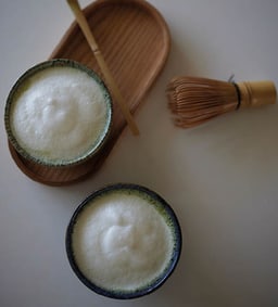 Zwei Schalen Matcha-Latte mit cremigem Milchschaum, daneben Bambusbesen und Tablett.