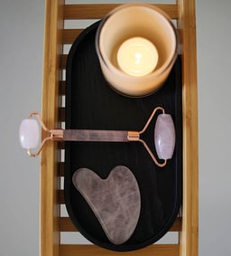 Rosenquarz-Roller und Gua-Sha-Stein auf einem schwarzen Tablett, daneben eine brennende Kerze.