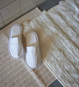 Weisse Badeslipper auf cremefarbenen Badteppichen im Badezimmer.
