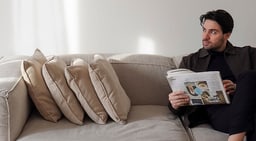 Personne assise sur un canapé lisant un magazine, entourée de coussins beiges.