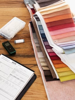 Stoffmuster in verschiedenen Farben, Nähzubehör und Formulare auf einem Holztisch.