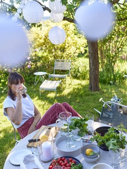 Una tavola apparecchiata in un giardino estivo con ingredienti freschi, candele e lanterne decorative sullo sfondo.