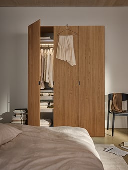 Offener Kleiderschrank aus Holz mit hängendem Rock, daneben ein Stuhl und ein Bett.