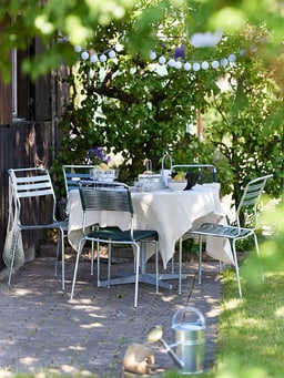 Due sedie in metallo accanto a un tavolo apparecchiato all&apos;ombra di un giardino, con una borsa a rete appesa allo schienale.