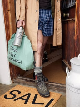 Personne en chaussures de randonnée et manteau avec un sac et une gourde sur un paillasson &quot;Sali&quot;.