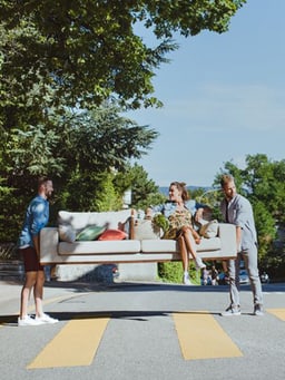 Du siehst zwei Personen, die ein Sofa im Freien tragen, auf dem eine Frau sitzt.